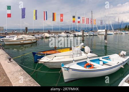 Kleine Boote und Yachten, die im Hafen von Bardolino vor Anker liegen, mit europäischen Flaggen an der Hafenmauer. Bardolino, Gardasee, Italien, Europa Stockfoto