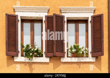 Nahaufnahme von 2 hübschen Fenstern mit Holzläden und Blumen in einem Gebäude mit goldgelben Wänden, Bardolino, Gardasee, Italien, Europa Stockfoto