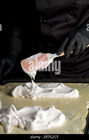 Der Konditor streut den Baiser mit einem kulinarischen Silikonspachtel auf dem gerollten Teig, um ihn im Ofen weiter zu backen Stockfoto