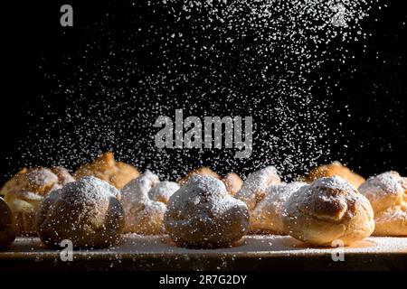 Gebackene Profiteroles mit Vanillesahne auf einer Holzplatte, die mit Puderzucker bestreut ist Stockfoto