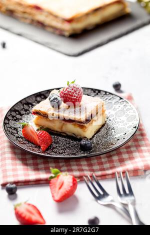 Ein Stück Napoleonkuchen mit Mohnsamen und Kirschen, dekoriert mit Erdbeeren und Blaubeeren auf hellem Hintergrund. Schließen Stockfoto