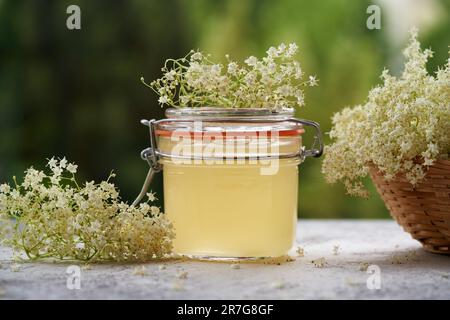 Ein Glas hausgemachter Holunderblütensirup mit frischen Blüten auf einem Tisch Stockfoto