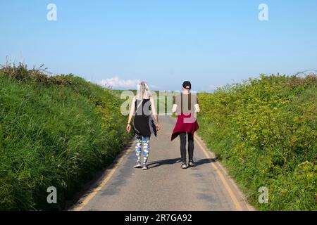 Rückansicht von zwei Freunden, die im Sommer an einem sonnigen Tag im Frühjahr 2023 auf einer Landstraße spazieren. Pembrokeshire Wales UK KATHY DEWITT Stockfoto