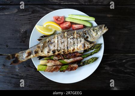 Gegrillter Seebarschfisch mit Zitronenstücken, frischer Salat und gegrillter Spargel mit Speck auf weißem Teller. Lebensmittelfotografie Stockfoto