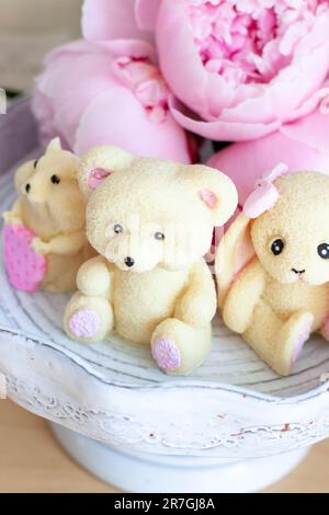 Süßes essbares Teddybär-, Häschen- und Hamsterspielzeug aus weißer Schokolade mit frischen Pfingstrosen und Kerzen im Hintergrund. Ein Geschenk für ein Baby Stockfoto