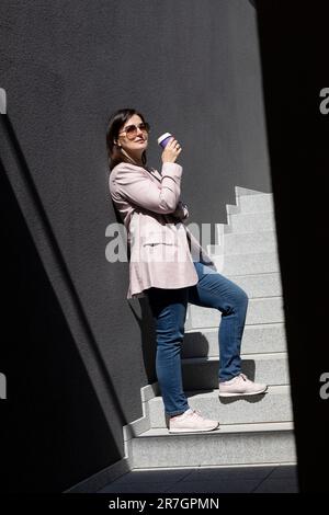 Stilvolle, Wunderschöne Brünette Frau Trinkt Kaffee Aus Dem Pappbecher, Steht Auf Der Treppe Neben Dem Bürogebäude Draußen, Sommer Sonniger Tag. Ich mache eine Pause Stockfoto