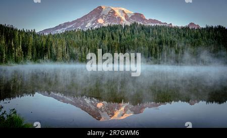 Mount Rainier und Kiefernwald spiegeln sich bei Sonnenaufgang in Reflection Lakes mit leichtem Nebel über dem See wider | Mount Rainier National Park, Washington, USA Stockfoto