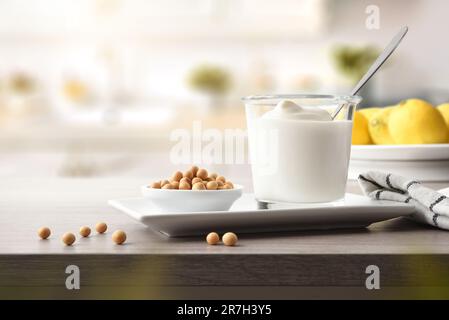 Glasbecher mit Sojamjoghurt und Schüssel mit Getreide auf Keramikplatte auf Holzbank in der Küche. Vorderansicht. Stockfoto