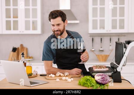 Mann mit einem Glas Wein beim Abendessen, während er sich den Online-Kochkurs mit einem Laptop in der Küche anschaute Stockfoto