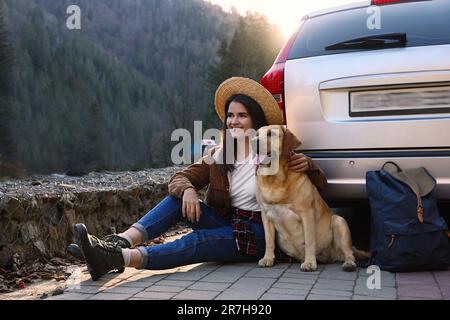 Eine glückliche Frau und ein niedlicher Hund, der in den Bergen neben dem Auto sitzt. Reisen mit Haustier Stockfoto