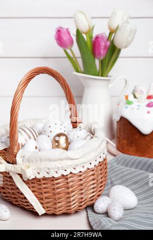 Korb aus Korb mit festlich dekorierten Eiern, wunderschönen Tulpen und traditionellem Osterkuchen auf weißem Holztisch Stockfoto