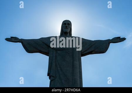 Vorderansicht der Christus-Erlöser-Statue mit dahinter verborgener Sonne auf dem Gipfel des Corcovado unter dem sonnigen, klaren blauen Himmel. Stockfoto