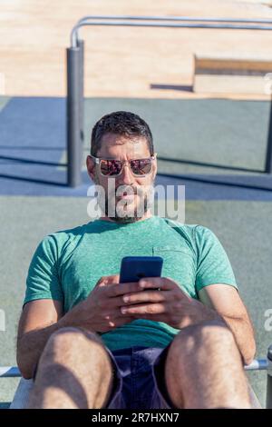 Ein junger Mann mit Bart und Sonnenbrille konsultiert eine Trainingsanwendung auf seinem Mobiltelefon, während er in einem Fitnessstudio im Park trainiert. Stockfoto