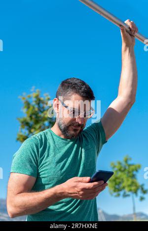 Ein junger Mann mit Bart und Sonnenbrille konsultiert eine Trainingsanwendung auf seinem Mobiltelefon, während er in einem Fitnessstudio im Park trainiert. Stockfoto