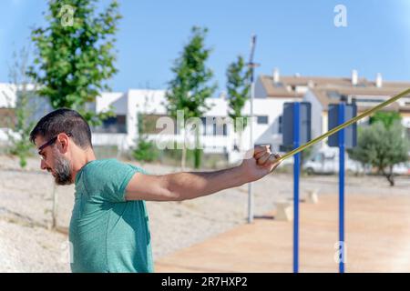 Profilansicht eines bärtigen Mannes mit einer Sonnenbrille, die seinen Rücken mit einem elastischen Riemen dehnt Stockfoto