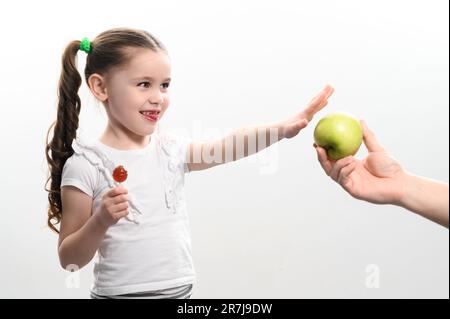 Das kleine Mädchen wählt einen Lutscher über einen Apfel, Süßigkeiten und Obst, die Wahl zwischen gesunder und ungesunder Nahrung, weißer Hintergrund und Kind. Stockfoto