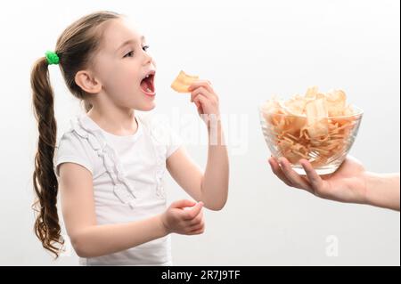 Das kleine Mädchen erhält eine große Schüssel Chips Snacks mit Schmalz, weißes Hintergrundporträt eines kleinen Mädchens, das Chips isst, ein Kind und ungesundes Fast Food. Stockfoto