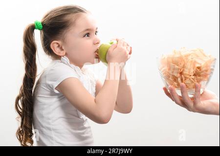 Große Schüssel mit Chips und Obst, wunderschönes kleines Mädchen isst einen Apfel, Kind bekommt eine Schüssel Chips, weißen Hintergrund und Kopierraum, gesund und Junk für Stockfoto