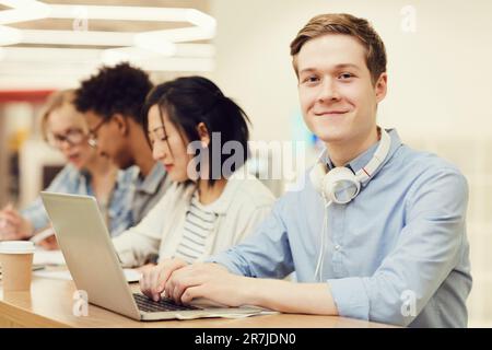 Porträt eines modernen jungen Schülers mit Kopfhörern am Hals, sitzt am Schreibtisch und tippt während des Vortrags auf dem Laptop Stockfoto