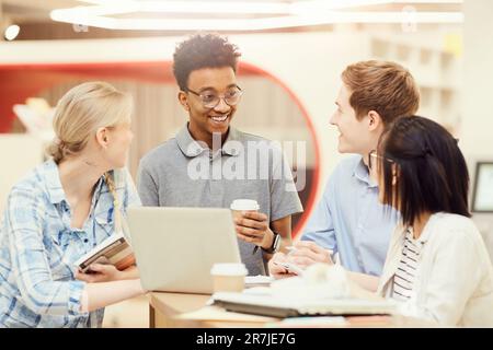 Eine Gruppe glücklicher junger interrassischer Schüler in lässigen Outfits, die mit einem Laptop in der Bibliothek an einem hohen Tisch stehen und Projektideen teilen Stockfoto