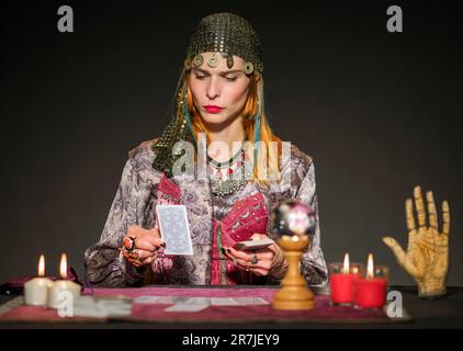 Fokussierter Wahrsager in authentischem Outfit und Münzkopfstück, der mit brennenden Kerzen am Tisch sitzt und mit Tarotkarten die Zukunft erraten kann Stockfoto