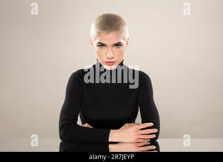 Porträt einer ernsten jungen Frau mit kurzem Haar im schwarzen Rollkragenpulli, die am Tisch sitzt und sich auf gekreuzte Hände lehnt, während sie gegen Lig in die Kamera schaut Stockfoto