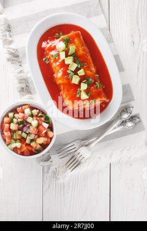 Mexikanische nasse Burrito-Platte mit roter Enchilada-Sauce, gekühlten Bohnen, Reis und Käse auf dem Teller auf dem Holztisch. Vertikale Draufsicht von Stockfoto