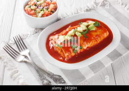 Leckere, nasse Burritos mit Rindfleisch, Reis und gekühlten Bohnen, verpackt in einer Tortilla, erstickt mit roter Sauce, auf dem Teller auf dem Holztisch. Stockfoto