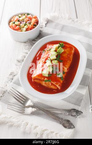 Mexikanische nasse Burrito-Platte mit roter Enchilada-Sauce, gekühlten Bohnen, Reis und Käse auf dem Teller auf dem Holztisch. Vertikal Stockfoto