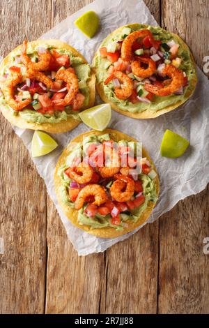 Köstliche Tostada mit Garnelen, frischem Gemüse und Guacamole aus nächster Nähe auf einem Holztisch. Vertikale Draufsicht von oben Stockfoto