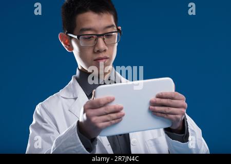 Asiatischer Wissenschaftler durchsucht Updates und Daten auf seinem Tablet. Ein chinesischer Ethnizitätsforscher vor blauem Hintergrund. Brille tragen, fasziniert von Sr Stockfoto