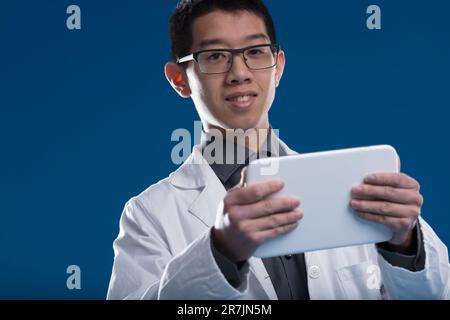 Ein Wissenschaftler chinesischer ethnischer Herkunft überprüft Daten auf seinem Tablet, fasziniert von Bildschirminhalten. Vor einem blauen Hintergrund, er ist mit Videos, Nachrichten, E beschäftigt Stockfoto