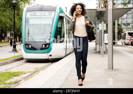Junge selbstbewusste Dame, die mit dem Smartphone auf einer Straßenbahnhaltestelle spaziert. Stockfoto