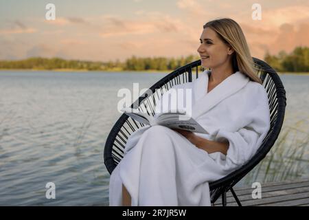 Frau im weißen Bademantel liest ein Buch im Stuhl auf der Fußgängerbrücke des Sees und genießt einen warmen Sommerabend Stockfoto