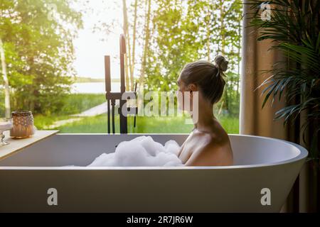 Junge Frau, die sich in der Badewanne voller Schaumstoff entspannt und aus dem Fenster im Badezimmer schaut Stockfoto