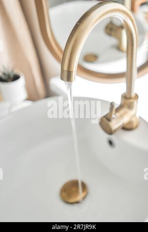 Ein wunderschönes Waschbecken mit angeschaltetem Wasser, goldenem Wasserhahn neben einem ovalen Spiegel und einem Regal mit Handtüchern. Nahaufnahme eines eleganten goldenen Wasserhahns Stockfoto