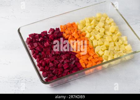 Rohes gewürfeltes Gemüse: Rüben, Karotten, mit Gewürzen gewürzte Kartoffeln und Pflanzenöl in einer Glasbackform auf hellgrauem Hintergrund Stockfoto