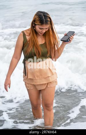 Halten Sie ihr Handy für Selfies, an einem Strand in der Nähe von Manila in der Dämmerung, schlendern Sie ziellos durch die warmen Küstengewässer, entlang des sanften Sandes, haben Sie Spaß, tanzen Sie herum Stockfoto
