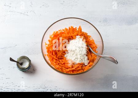 Glasschüssel mit geriebenen Karotten mit Mehl und Salz auf einem hellgrauen Tisch, Draufsicht. Hausgemachte vegetarische Speisen: Karotten-Pfannkuchen, Pommes Frites oder Stockfoto
