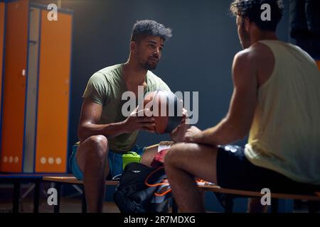 Zwei gutaussehende junge Basketballspieler sitzen in der Garderobe auf einer Bank und unterhalten sich nach dem Training. Aktiver Lifestyle-Konz Stockfoto