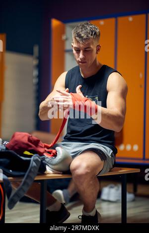 Attraktiver junger Mann sitzt in der Garderobe, konzentriert sich, während er sich zum Schutz mit Klebeband um die Hand bindet, und bereitet sich auf das Boxtraining vor. Stockfoto