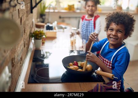 Afro-amerikanische Geschwister freuen sich auf kulinarische Entdeckungsreise. Der kleine Junge gibt verspielt vor, Gemüse in einer Pfanne zu kochen. Im Hintergrund Stockfoto