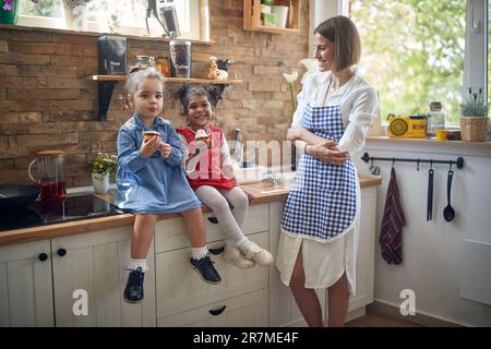 Fröhliche junge Mutter zusammen in der Küche mit zwei kleinen Mädchen, die auf der Küchentheke sitzen, Muffins essen und sich fröhlich fühlen. Zuhause, Familie, Stockfoto