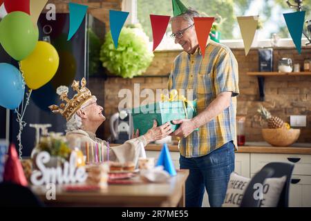 Fröhliche Seniorin, die von ihrem Mann ein Geschenk erhält, glücklich ist, in der Küche sitzt und aufblasbare goldene Krone trägt. Lebensstil, Seniorenleben, Feiern Stockfoto