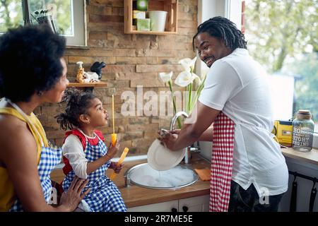 Die afro-amerikanische Familie teilt fröhliche Momente in ihrer Küche. Der Vater wäscht das Geschirr, während die Mutter ihr niedliches kleines Mädchen liebevoll festhält Stockfoto