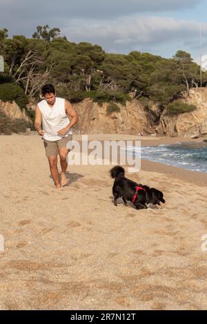 Ein Mann mit seinem Hund läuft am Strand mit üppigen Bäumen im Hintergrund. Stockfoto