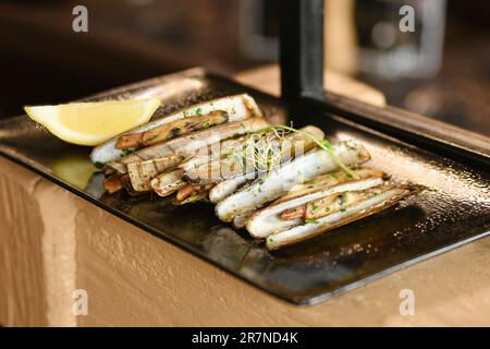 Die Muschelmesser, Petersilie und eine halbe Zitrone auf schwarzem Teller Stockfoto