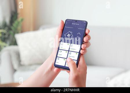 Die Hand einer Frau hält ein Mobiltelefon und zeigt eine Smart-Home-App. Smart Living-Konzept. Dekoriertes Wohnzimmer im Hintergrund Stockfoto