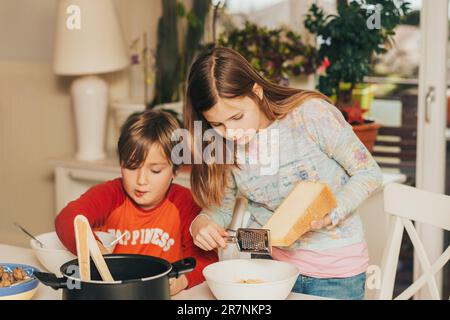 Zwei Kinder essen zu Hause Spaghetti, ein Mädchen streut Nudeln mit einem großen Stück Käse Stockfoto