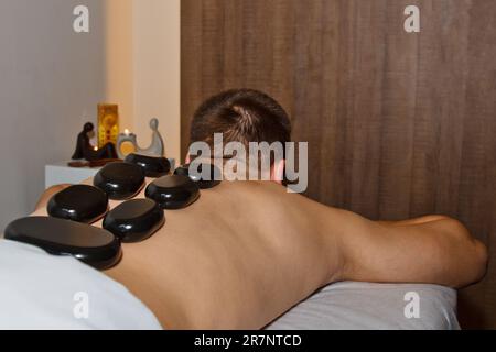 Wellnesskonzept. Attraktiver junger Mann, der sich unter dem stimulierenden Effekt einer traditionellen Hot-Stone-Massage in einem luxuriösen Spa und Wellnesscenter entspannen kann. Aufn Stockfoto